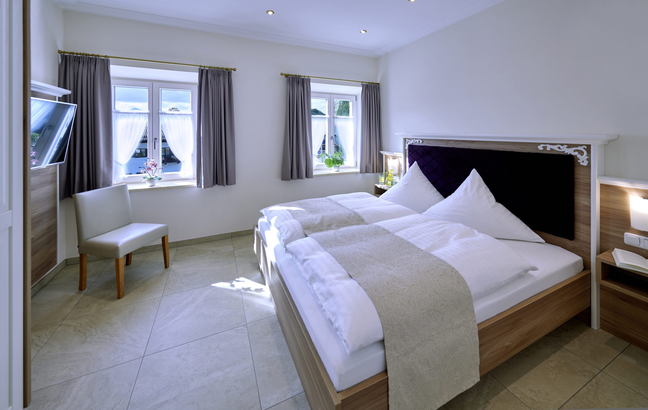 schlafzimmer mit hellbraunem doppelbett, am kopfteil ist der mittlere bereich mit lila samt verkleidet und die ecken mit weissen ornamenten verziert