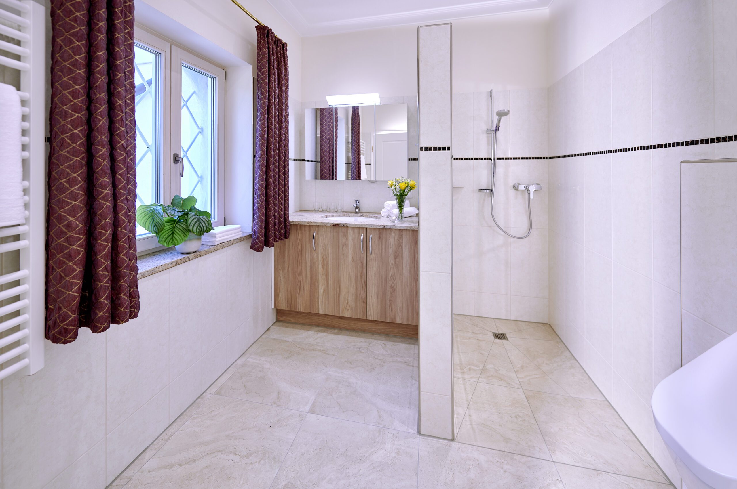 badezimmer mit begehbarer dusche auf der rechten seite, links daneben ist der waschtisch mit beige-melierten granit und hellbraunen unterbauschrank, am fenster sind rote vorhaeinge